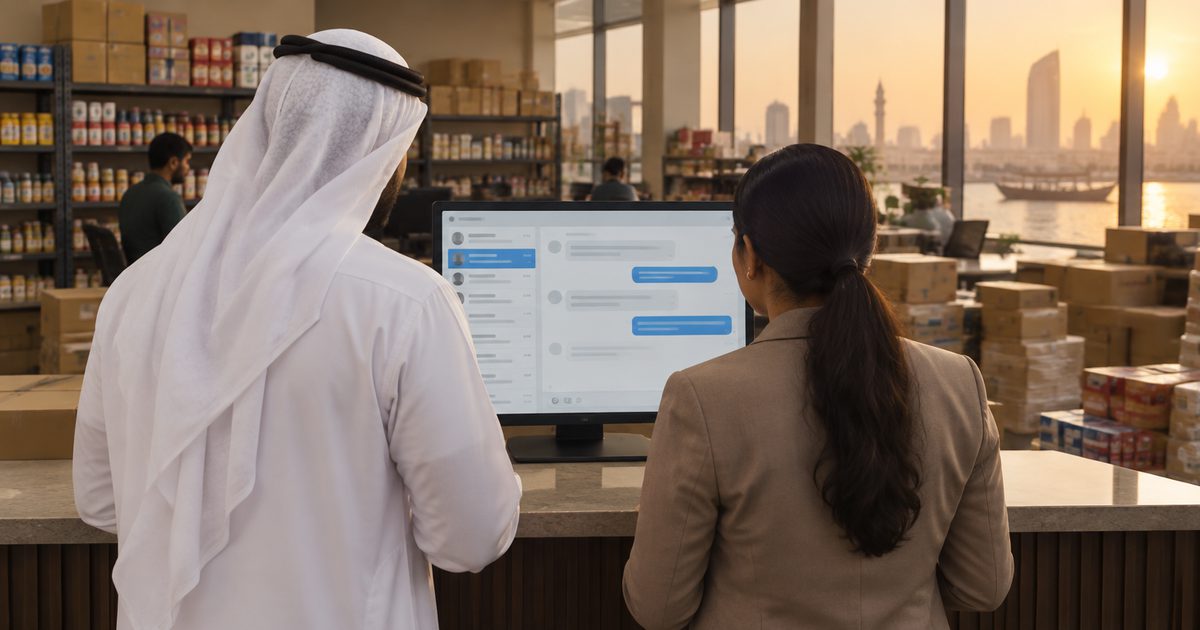 Two staff at a Deira trading showroom counter, seen from behind, reviewing a WhatsApp support inbox on a monitor, with stocked shelves and Dubai Creek behind them at sunset